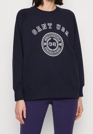 Kobieta w granatowej bluzie z napisem „GANT USA” i białym, okrągłym logo „American Sportswear 1949”, w połączeniu z fioletowymi spodniami.