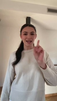 Mladá žena s tmavými vlasmi v bielom svetri gestikuluje zdvihnutým ukazovákom a hovorí v interiéri pred jednoduchou béžovou stenou.