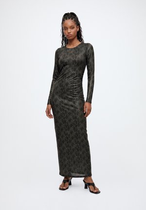 Femme aux cheveux tressés portant une robe longue à manches longues avec un imprimé serpent sombre et des sandales noires à talons, debout sur fond blanc.