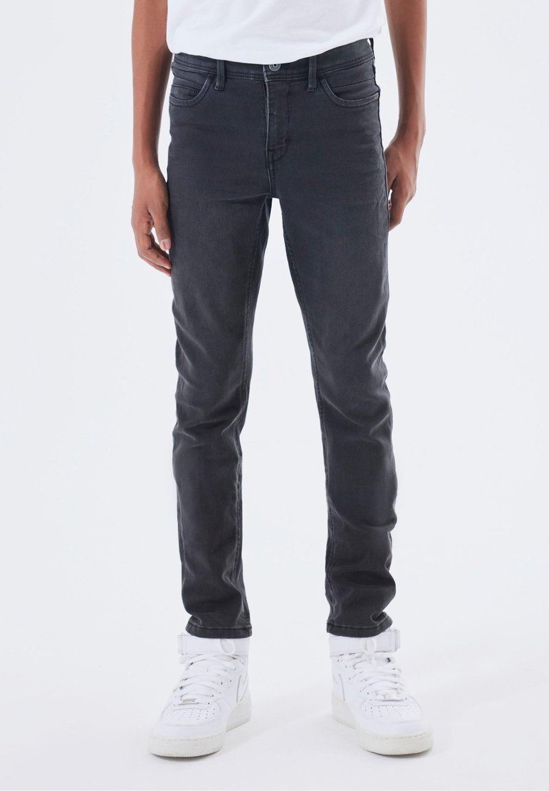 Personne portant un jean slim gris foncé et des baskets montantes blanches, debout devant un fond uni.