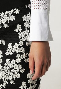 Czarna spódnica z białymi haftami kwiatowymi, teksturowany materiał, zestawiona z białą bluzką z ażurowymi detalami na rękawach.