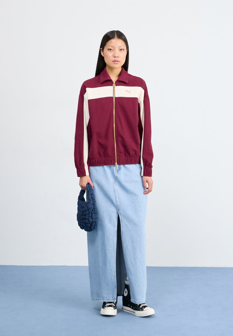 Bordeauxfarbene und cremefarbene Zip-Jacke mit goldfarbenen Details, kombiniert mit einem hellblauen Denim-Rock mit einem Schlitz vorne. Hält eine dunkelblaue gehäkelte Tasche.