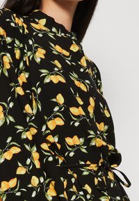 Primo piano di un indumento nero con collo alto, vita cinte con una cintura, e motivo di frutti giallo-arancio brillanti e foglie verdi.