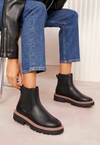 Botas de piel negra hasta el tobillo con paneles laterales elásticos, puntera redonda y suela gruesa marrón; combinadas con jeans cortos azules.