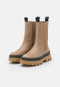 Bottines beige avec des semelles épaisses couleur tan, des bordures vertes, des panneaux latéraux élastiques et des poignées noires sur un fond blanc.