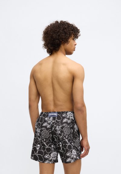 Mladý muž s kučeravými vlasmi, odhalený chrbát otočený preč, oblečený v čiernych plavkách s bielymi kvetinovými vzormi na jednofarebnom pozadí.
