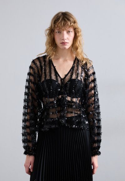 Black sheer blouse with a textured sequin pattern, long sleeves, and a front tie detail, paired with a black pleated skirt.