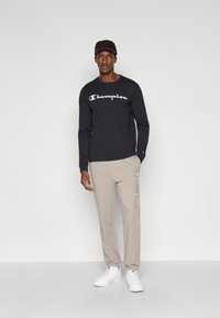 Maglietta nera a maniche lunghe con logo "Champion" bianco, pantaloni della tuta beige e scarpe da ginnastica bianche. Modello con cappellino nero. Design semplice e casual.