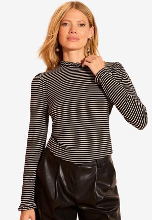 Femeie purtând un top cu mâneci lungi, în dungi alb-negru, cu guler și manșete cu volane, asortat cu pantaloni negri din piele, pozând cu mâna lângă față.