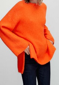 Pull orange vif surdimensionné, tricot texturé, encolure ronde et épaules tombantes. Présente des fentes sur les côtés et des poignets côtelés.
