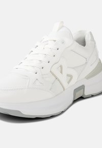 Weißer Sportschuh mit strukturiertem Obermaterial, Mesh-Akzenten, Schnürverschluss und einer dicken weißen Sohle mit grauem Akzent in der Nähe der Ferse.