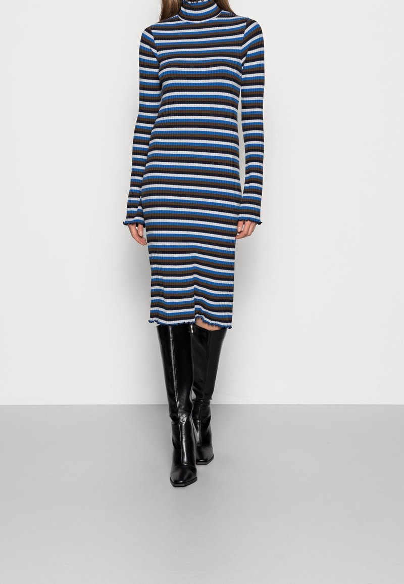 Ribbade stickade klänningen med svarta, blåa och bruna horisontella ränder; hög krage, långa ärmar och rå nederkant; tillsammans med knähöga svarta stövlar.