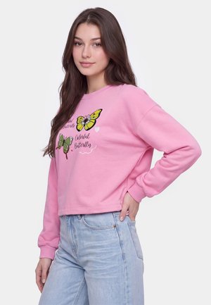 Ung kvinde iført en lyserød sweatshirt med gule og grønne sommerfuglemotiver og lyseblå jeans, poserer med den ene hånd på hoften.