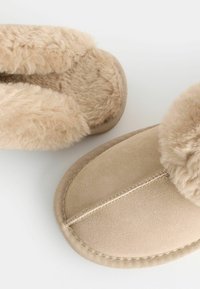 Pantofole beige con un morbido interno in peluche, esterno in suede, punta rotonda e dettagli ricamati lungo la suola.