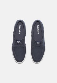 Navyfarbene Canvas-Slipper mit runder Zehenpartie, weißer Gummisohle, Schnürdetails und Timberland-Logo an der Seite sowie auf der Innensohle.