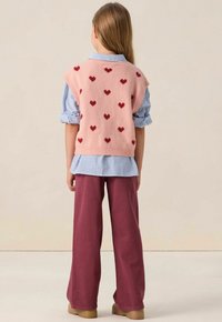 Une jeune personne de dos, portant un gilet rose avec des cœurs rouges, une chemise rayée bleue, un pantalon bordeaux et des chaussures beiges.