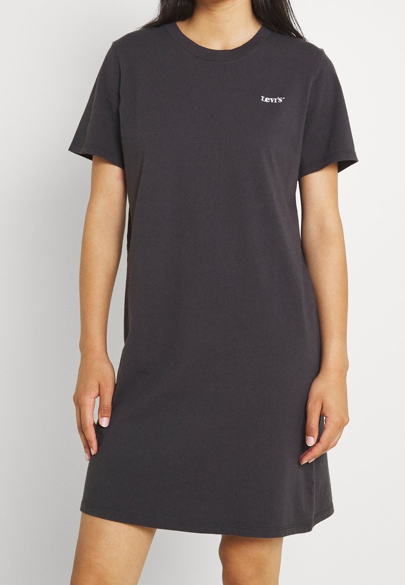Personne portant une robe T-shirt noire Levi's à manches courtes, longueur genou, avec un logo sur la poitrine gauche, sur fond uni.
