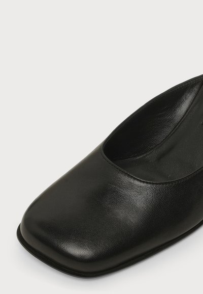 Chaussure plate en cuir noir avec un bout carré et une finition lisse, montrée en gros plan sur un fond blanc.