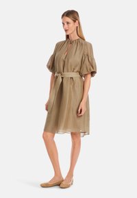 Khakifarbenes Leinenkleid mit hohem Kragen, Puffärmeln, tailliertem Gürtel und knielanger Hemdlänge. Kombiniert mit gewebten beigen Ballerinas.