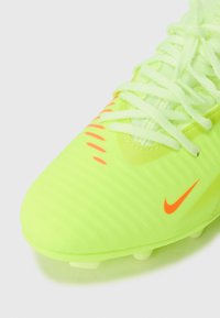 Bota de fútbol de un brillante color verde neón con el logo y acentos naranjas de Nike, con un empeine texturizado y cordones blancos, en ángulo sobre un fondo blanco.