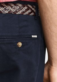 Lähikuva laivaston sinisten housujen takataskusta, jossa on nappi ja GANT-label, puettuna punotun ruskean ja beigen vyön kanssa, paljaan käden vieressä.