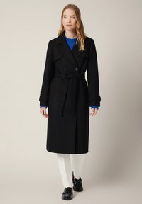 Czarny podwójnie zapinany trencz z paskiem w talii i szerokimi klapami, połączony z niebieskim swetrem i białymi spodniami, wykończony czarnymi butami.