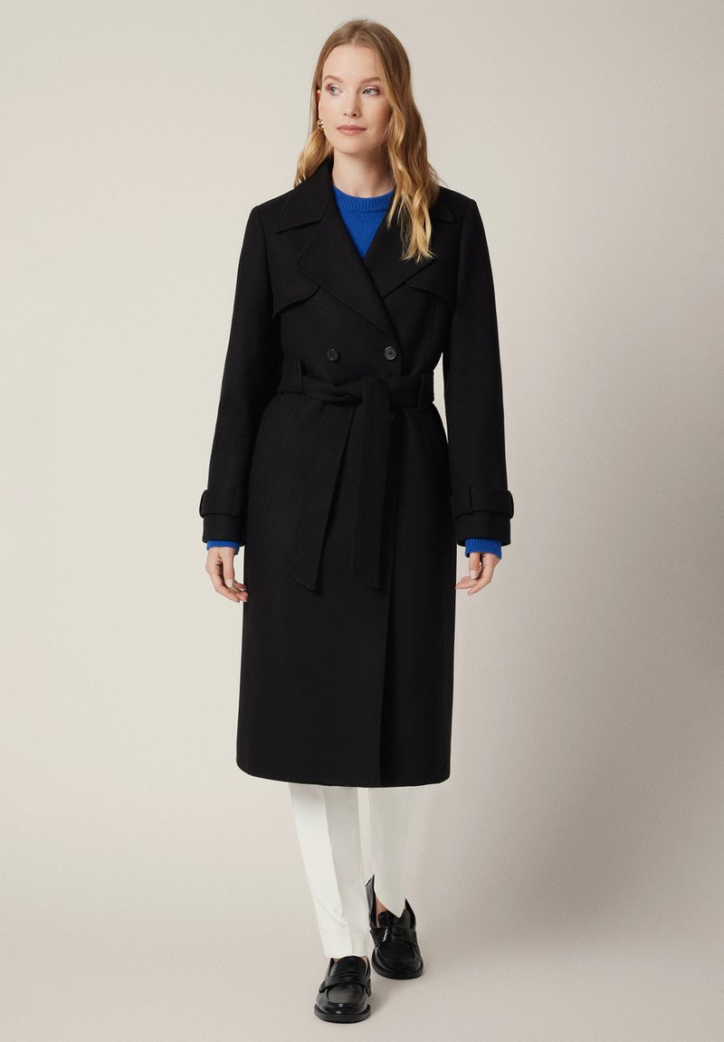 Czarny podwójnie zapinany trencz z paskiem w talii i szerokimi klapami, połączony z niebieskim swetrem i białymi spodniami, wykończony czarnymi butami.