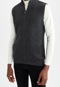 Gilet lavorato a maglia grigio scuro con colletto alto, zip frontale e due tasche laterali. Indossato sopra una camicia bianca a maniche lunghe, in tessuto texturizzato.