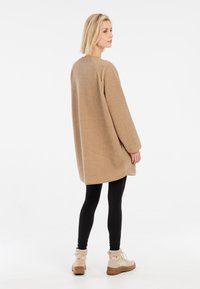 Beige fleece oversized jas met lange mouwen, ronde hals en afgeronde zoom, gecombineerd met zwarte leggings en beige enkellaarsjes.