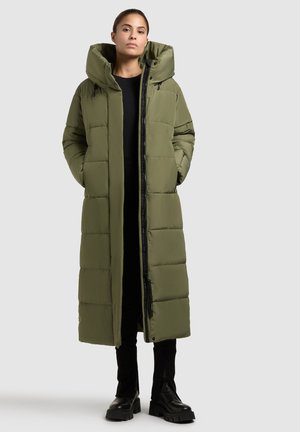 Dlhý olivovo zelený puffový kabát s vysokým golierom, predným zipsom, bočnými vreckami a prešívaným dizajnom. Model má na sebe čierny top a nohavice.