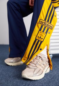 Scarpe da atletica beige con pannelli testurizzati e accenti bianchi, abbinate a pantaloni da jogging blu navy e giallo con tre strisce e una toppa con logo.