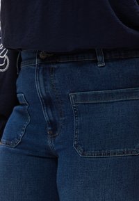 Σκούρο μπλε τζιν με ορατές πίσω τσέπες και κλείσιμο με κουμπί, σε συνδυασμό με σκούρο μπλε τοπ με λευκή κεντητική στο μανίκι.