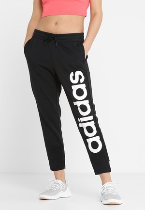 Osoba nosící černé tepláky Adidas s velkým bílým logem na levé noze, doplněné bílými teniskami a korálovým krátkým topem.