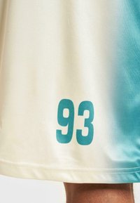 Šortky s prechodom farieb od smotany po svetlo modrú, s veľkým tyrkysovým potlačením čísla "93" na leme, vyrobené z hladkej látky s plochými švami.