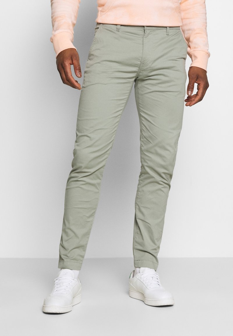 Light green, slim-fit chinos made of smooth fabric, featuring side pockets and a zip fly, paired with white sneakers.