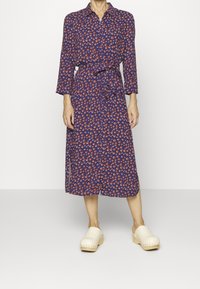 Vestito midi blu con motivi circolari rossi e bianchi, apertura con bottoni, colletto e vita aderente; abbinato a zoccoli crema su uno sfondo neutro.