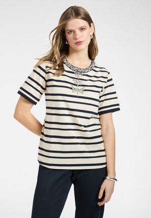 Mujer con cabello lacio castaño claro, vistiendo una blusa de manga corta a rayas en blanco y negro con joyas decorativas y pantalones oscuros, posando contra un fondo blanco.