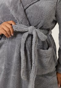 Grå, fluffig badrock med en mjuk textur, med bälte och två framfickor. Tyget verkar varmt och bekvämt.