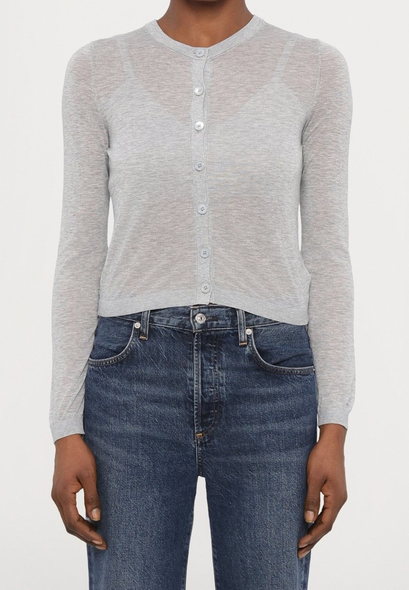 Femme portant un cardigan gris clair boutonné et un jean taille haute bleu foncé, debout avec les bras relâchés le long du corps.
