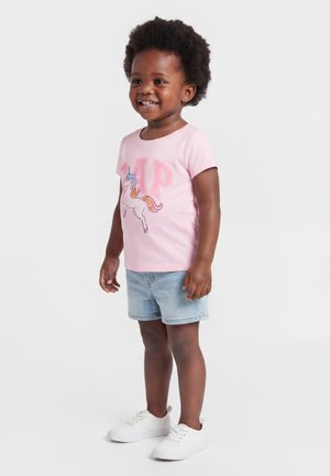Mosolygó kisgyerek rózsaszín unikornis mintás pólóban, világos farmer rövidnadrágban és fehér sportcipőben, fehér háttér előtt áll.