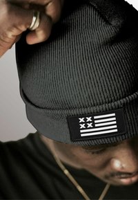 Black ribbed beanie with a folded cuff, featuring a rectangular patch displaying white stripes and two black Xs. Soft texture.