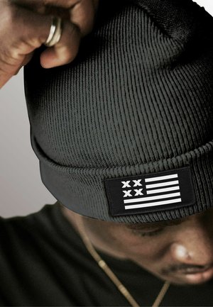 Black ribbed beanie with a folded cuff, featuring a rectangular patch displaying white stripes and two black Xs. Soft texture.