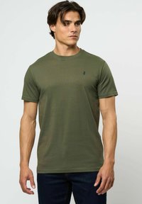 Vīrietis ar tumšiem matiem, valkājot olīvu zaļu apaļu kakla T-kreklu un tumši zilas džinsas, skatās uz sāniem pret vienkrāsainu gaišu fonu.