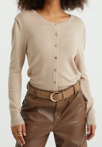 Kvinna som bär en beige knäppt cardigan med guldknappar, bruna läderbyxor och ett matchande brunt bälte med en guldspänne.