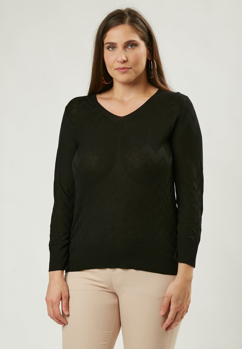 Schwarzer V-Ausschnitt-Pullover mit langen Ärmeln und Zick-Zack-Muster; aus leichtem Strickstoff; gerippte Bündchen und Saum. Kombiniert mit beigen Hosen.