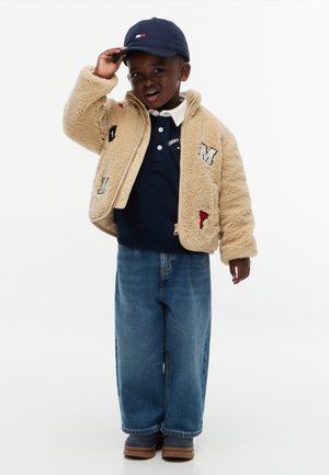 Jeune enfant portant une casquette bleu marine, une veste beige pelucheuse avec des patches, une chemise bleu marine, un jean large bleu et des chaussures marron, debout et touchant la casquette.