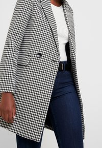 Cappotto a fantasia houndstooth bianco e nero con rever a punta, chiusura con un bottone e due tasche frontali. Indossato sopra una maglia bianca a coste.