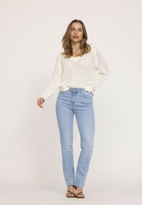 Pull en tricot blanc à mailles ouvertes avec un col en V, associé à un jean slim bleu clair et des sandales à talons hauts marron. Tenue casual minimaliste.