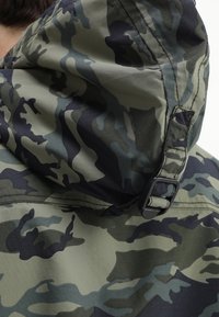 Giacca camouflage con cappuccio; presenta un motivo verde e nero, una texture liscia e una cinghia laterale con dettagli in metallo.