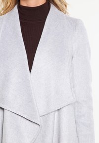 Cappotto in misto lana grigio con colletto drappeggiato, texture morbida e design aperto, indossato sopra un dolcevita marrone a coste.